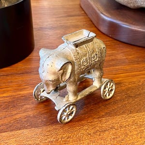 May include: A small, antique-style elephant figurine on wheels. The elephant is painted in a distressed, off-white colour with detailed carvings on its body. The wheels are also off-white and have a spoke design. The figurine is a decorative item.