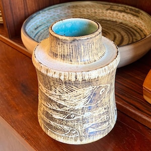 May include: A ceramic vase with a cylindrical shape and a light brown and beige color scheme. The vase features a textured surface with etched fish designs and a turquoise interior. The vase is displayed on a wooden surface.