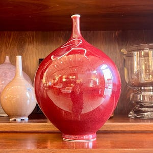 May include: A glossy, red ceramic vase with a bulbous body and a slender neck. The vase features a white, abstract design and sits on a wooden shelf. Other decorative vases are visible in the background.