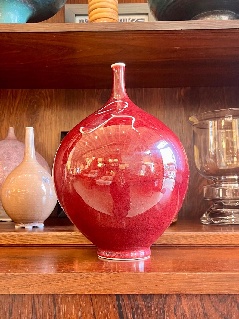 May include: A glossy, red ceramic vase with a bulbous body and a slender neck. The vase features a white, abstract design and sits on a wooden shelf. Other decorative vases are visible in the background.