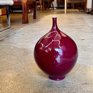 May include: A large, glossy, burgundy-colored vase with a rounded body and a slender neck. The vase has a white accent design and sits on a small, circular base. The vase is a decorative home decor item.