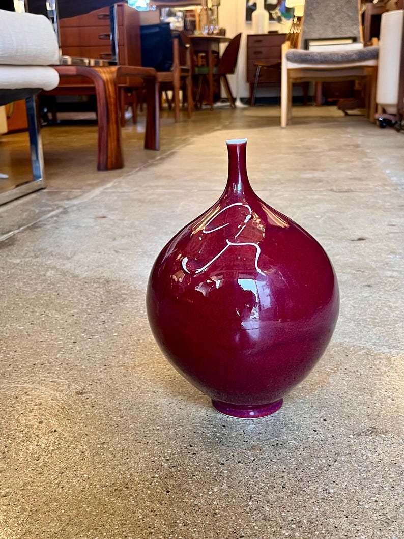 May include: A large, glossy, burgundy-colored vase with a rounded body and a slender neck. The vase has a white accent design and sits on a small, circular base. The vase is a decorative home decor item.