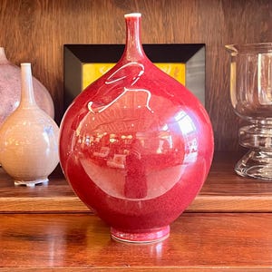 May include: A glossy, red decorative vase with a bulbous body and a slender neck. The vase has a white accent and sits on a wooden shelf. Other vases are visible in the background, suggesting a collection of decorative items.