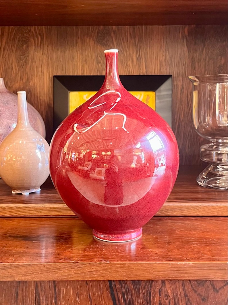 May include: A glossy, red decorative vase with a bulbous body and a slender neck. The vase has a white accent and sits on a wooden shelf. Other vases are visible in the background, suggesting a collection of decorative items.