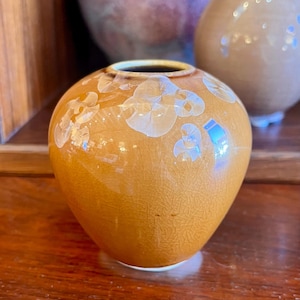 May include: A small, round, orange ceramic vase with a glossy finish. The vase features a crackled texture and white, floral-like patterns. The opening is dark brown. Two other vases are visible in the background.