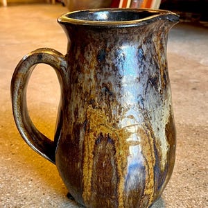 May include: A handcrafted ceramic pitcher with a dark brown, tan, and gold glaze. The pitcher has a handle and a spout for pouring. The glaze has a dripped effect, adding to its rustic charm. This decorative pitcher is perfect for serving beverages.
