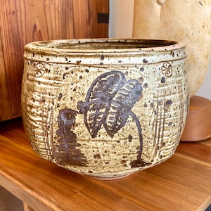 Puede incluir: Jardinera de cerámica hecha a mano con acabado moteado beige y marrón. La jardinera presenta un diseño de hojas marrón oscuro y patrones decorativos alrededor del borde. Ideal para uso en interiores o exteriores.