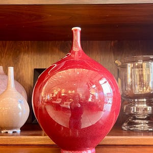 May include: A glossy, red ceramic vase with a bulbous body and a slender neck. The vase has a smooth, reflective surface, and sits on a wooden shelf. The vase is a decorative item, and is part of a collection of vases and bowls.