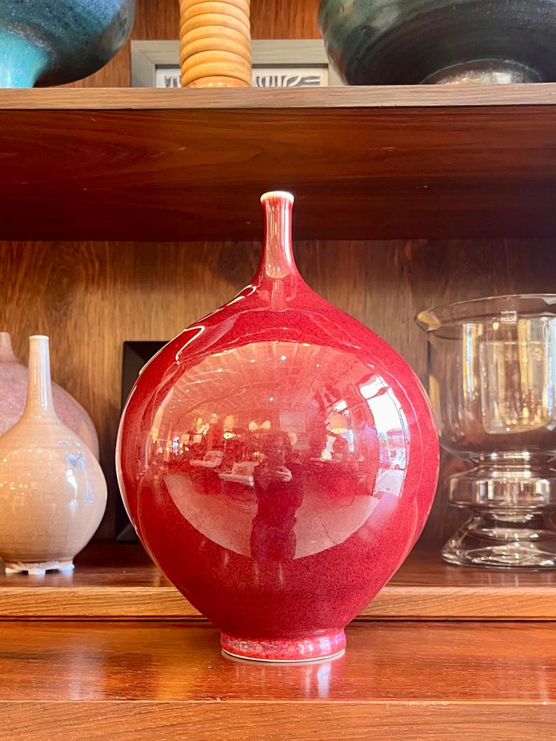 May include: A glossy, red ceramic vase with a bulbous body and a slender neck. The vase has a smooth, reflective surface, and sits on a wooden shelf. The vase is a decorative item, and is part of a collection of vases and bowls.