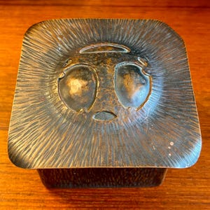 May include: A square, copper box with a hammered finish. The lid features a raised, stylized face with two large eyes and a small mouth.