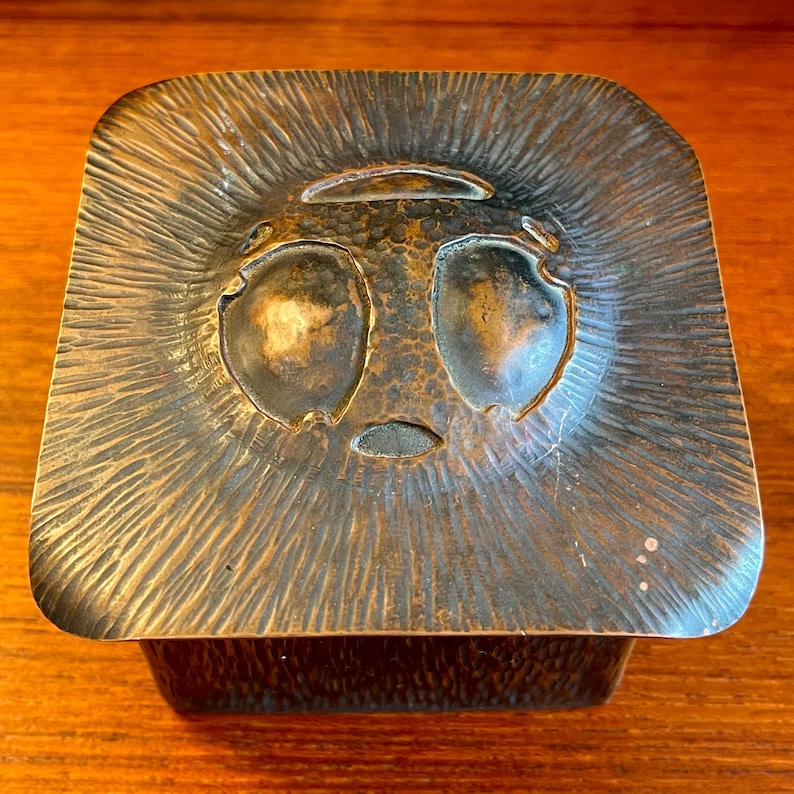 May include: A square, copper box with a hammered finish. The lid features a raised, stylized face with two large eyes and a small mouth.