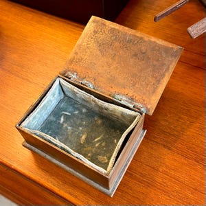 May include: A small, rectangular, copper box with a hinged lid. The box is open and the inside is lined with a light gray fabric.