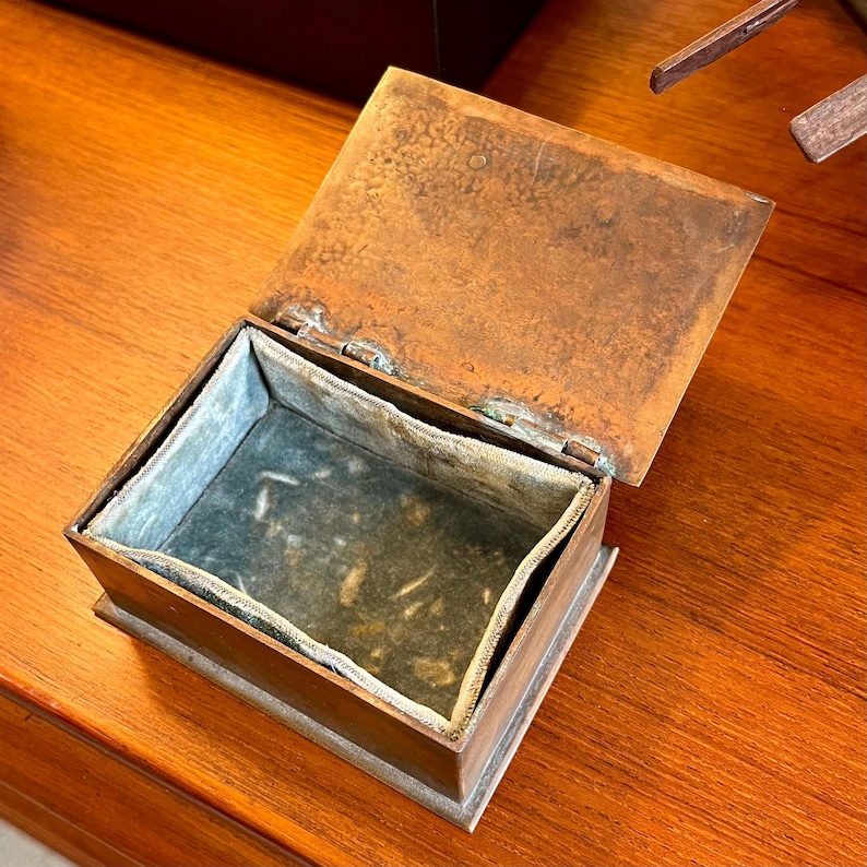May include: A small, rectangular, copper box with a hinged lid. The box is open and the inside is lined with a light gray fabric.