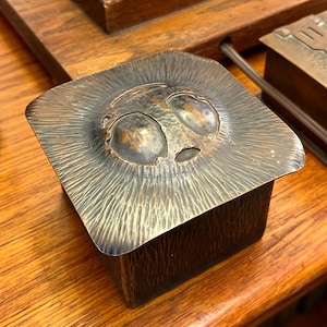 May include: A square, copper box with a hammered finish. The lid features a raised, stylized design of two eyes and a mouth.