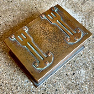 May include: A hammered copper box with a geometric design. The box has a rectangular shape and a textured surface. The design is in a dark green color.