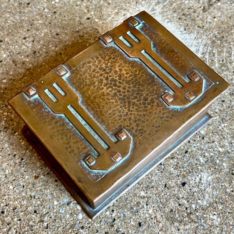 May include: A hammered copper box with a geometric design. The box has a rectangular shape and a textured surface. The design is in a dark green color.