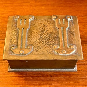May include: A rectangular copper box with a hammered finish and geometric designs. The box has a hinged lid and a raised edge.