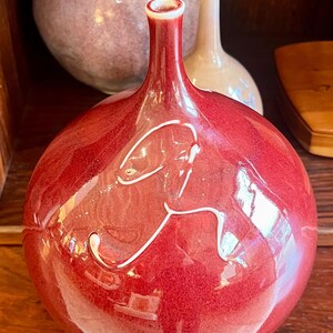 May include: A glossy, red ceramic vase with a bulbous body and a slender neck. The vase features a white, abstract design and a white rim. Other ceramic vases are visible in the background. The vase is likely used for decorative purposes.