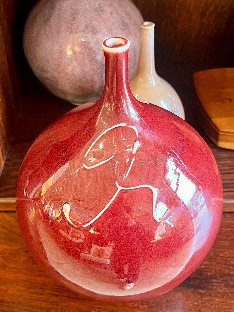 May include: A glossy, red ceramic vase with a bulbous body and a slender neck. The vase features a white, abstract design and a white rim. Other ceramic vases are visible in the background. The vase is likely used for decorative purposes.