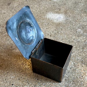 May include: A small, square, copper box with a hinged lid. The lid has a raised, circular design in the center. The box is made of hammered copper and has a rustic, aged look.