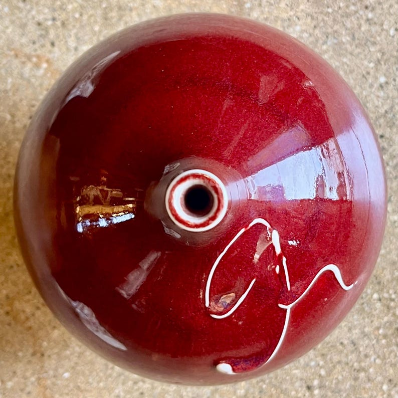 May include: A glossy, deep red ceramic object with a circular shape. It features a central opening with a white and brown ring and a white, stylized design on the side. The object has a smooth, reflective surface.