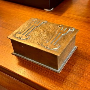 May include: A brass box with a hammered finish and an intricate geometric design on the lid. The box is closed and sitting on a wooden surface.