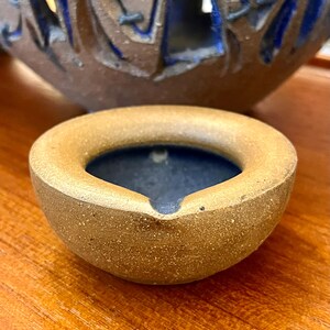 May include: A small, round, ceramic ashtray with a brown exterior and a dark blue interior. The ashtray has a thick, rounded rim and a small notch on one side. A larger, decorative ceramic piece is visible in the background.