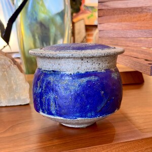 Può includere: Un vaso in ceramica fatto a mano con coperchio. Il vaso presenta una vivace glassa blu sul corpo, contrastata da un bordo e un coperchio grigio chiaro. La consistenza della glassa è visibile, aggiungendo al tocco artigianale. Il vaso è appoggiato su una superficie di legno.