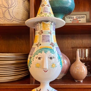 May include: A whimsical ceramic vase with a face design. The vase features a white base, a yellow, blue, and green patterned body, and a hat-like top. The face has blue hair, a smiling mouth, and yellow floral accents. The vase is displayed on a wooden shelf.