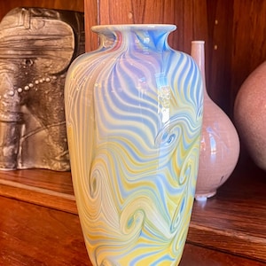 Puede incluir: Un jarrón de vidrio decorativo con un patrón arremolinado de amarillo, azul y blanco. El jarrón tiene una base ancha y un cuello estrecho. Otros jarrones son visibles en el fondo.