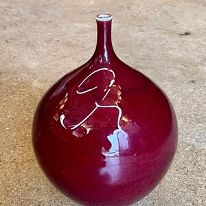 May include: A burgundy-colored ceramic vase with a bulbous body and a slender neck. The vase has a glossy finish and a white, abstract design on the front. The base and rim have a lighter shade of burgundy.