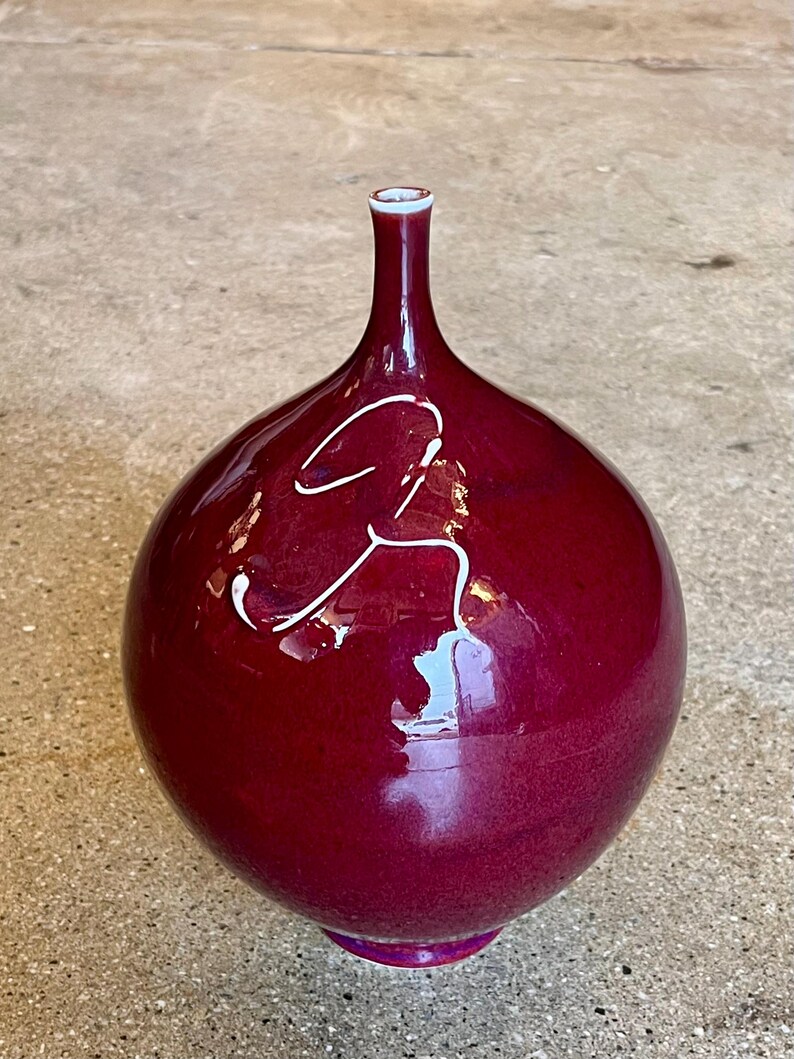 May include: A burgundy-colored ceramic vase with a bulbous body and a slender neck. The vase has a glossy finish and a white, abstract design on the front. The base and rim have a lighter shade of burgundy.
