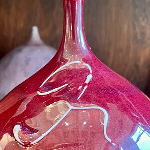 May include: A close-up of a red glass vase with a white rim and white abstract design. The vase has a long neck and a rounded body. Another vase is slightly visible in the background.
