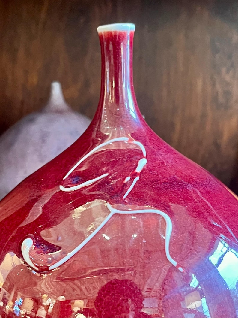 May include: A close-up of a red glass vase with a white rim and white abstract design. The vase has a long neck and a rounded body. Another vase is slightly visible in the background.