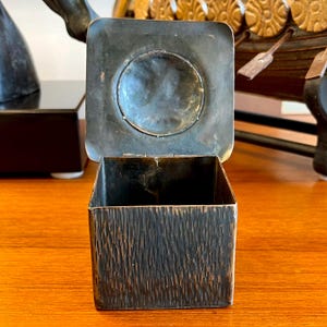May include: A square, dark brown, metal box with a textured surface and a hinged lid. The lid has a circular, recessed area in the center.