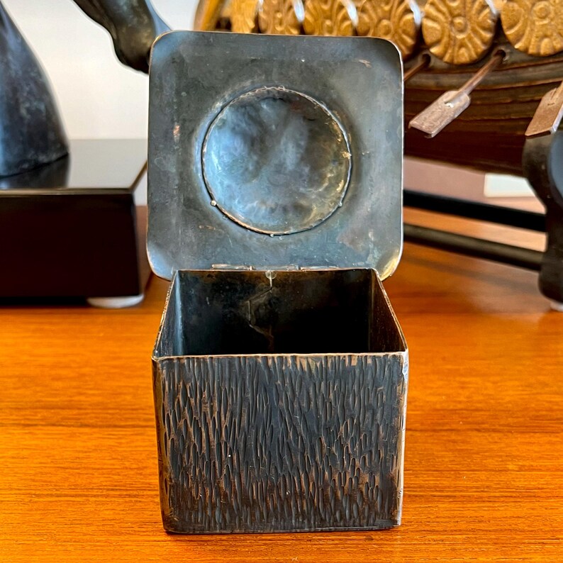 May include: A square, dark brown, metal box with a textured surface and a hinged lid. The lid has a circular, recessed area in the center.