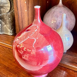 May include: A collection of decorative ceramic vases. The primary vase is a vibrant red with a glossy finish and white abstract detailing. Two other vases are visible in the background, one light pink and the other beige.