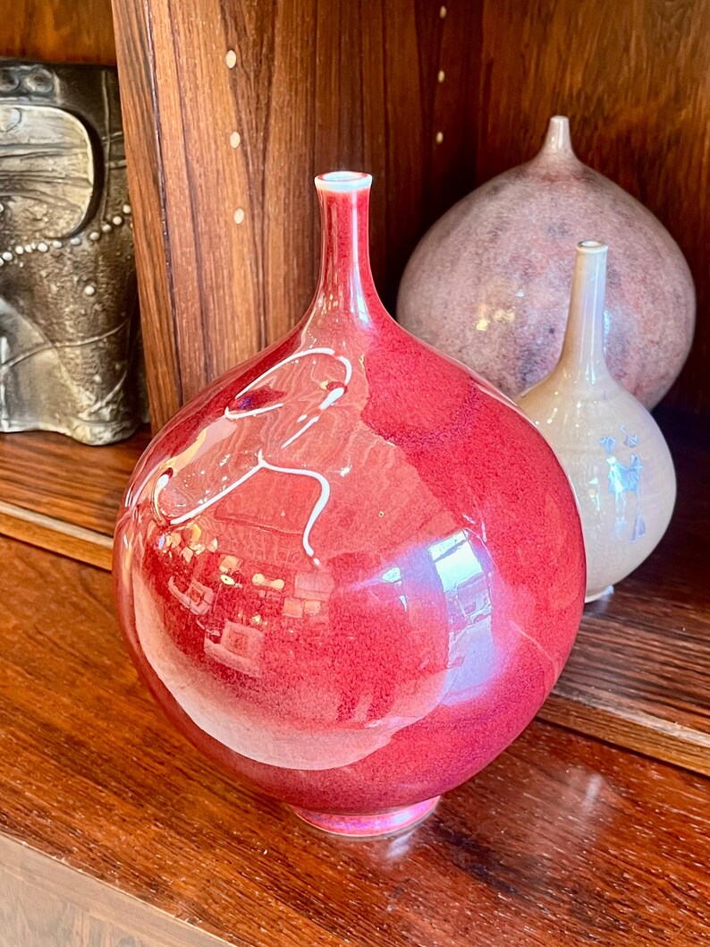 May include: A collection of decorative ceramic vases. The primary vase is a vibrant red with a glossy finish and white abstract detailing. Two other vases are visible in the background, one light pink and the other beige.