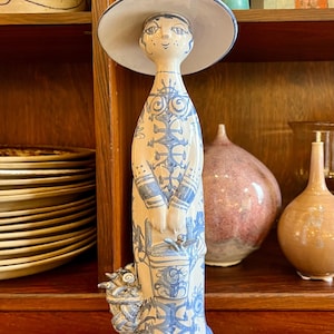 Może przedstawiać: Ceramiczna figurka kobiety w kremowo-niebieskich barwach, noszącej kapelusz z szerokim rondem. Figurka ma szczegółową twarz i jest ozdobiona niebieskimi wzorami kwiatowymi. Trzyma kosz kwiatów i jest przedmiotem dekoracyjnym.