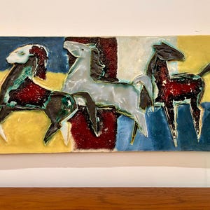 Może przedstawiać: Ceramiczna dekoracja ścienna przedstawiająca trzy stylizowane konie w odcieniach czerni, bieli i czerwieni. Konie umieszczono na tle żółtych, niebieskich i czerwonych figur geometrycznych. Praca jest podpisana "G.245".