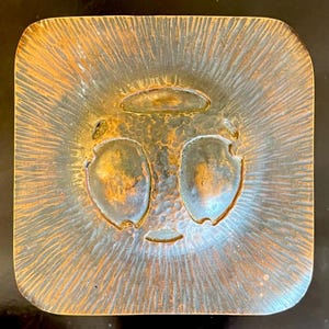 May include: A square, hammered copper tray with a raised, textured design. The tray has a circular, recessed area in the center with two smaller, rounded indentations.