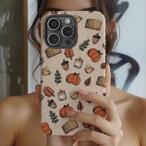 May include: A beige phone case with a fall-themed design. The case features pumpkins, acorns, leaves, coffee cups, and books. The case is designed for an iPhone with a triple lens camera.