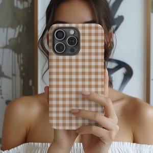 Op de afbeelding: Een bruin en wit geruit telefoonhoesje. De hoes wordt voor het gezicht van een persoon gehouden, waardoor het gezicht wordt bedekt. De telefoonhoes heeft een camerabult met drie lenzen.