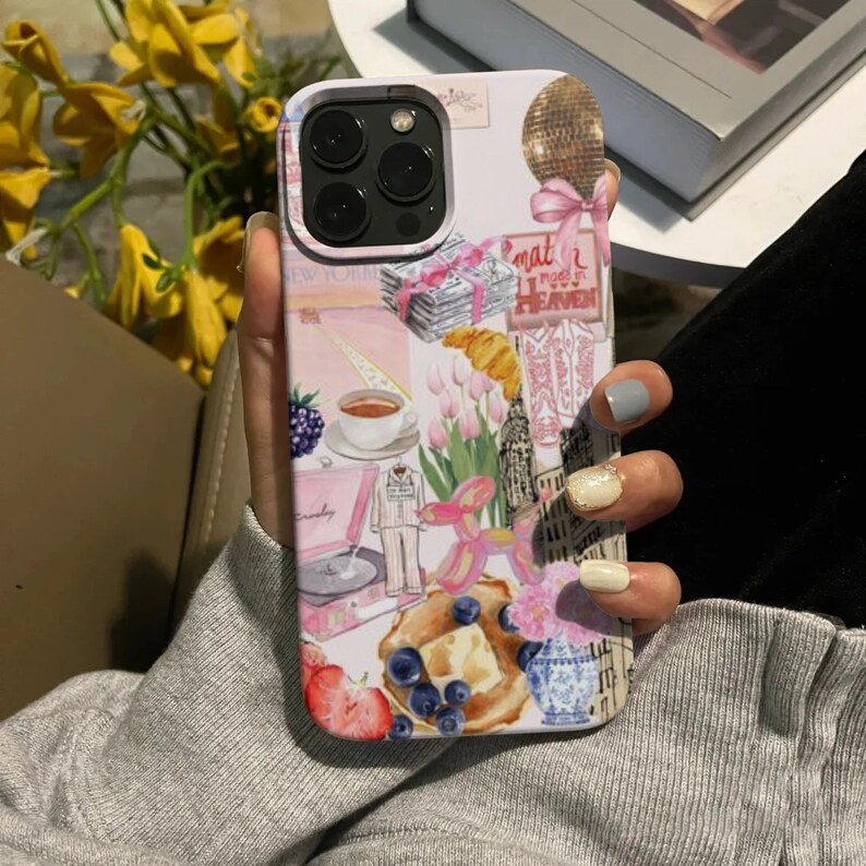 Op de afbeelding: Een roze iPhone-hoesje met een collage-ontwerp met een discobal, een platenspeler, een kopje koffie, een croissant, een bos tulpen, een ballondier en een aardbei. De hoes heeft ook de tekst "What I need in Heaven".