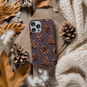 May include: A purple phone case with a pattern of brown pine cones. The case is on a wooden surface with fall leaves and a white knitted blanket.