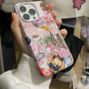 Op de afbeelding: Een roze en witte iPhone-hoes met een collage van afbeeldingen, waaronder een discobal, een kopje koffie, een platenspeler, een croissant en een bos tulpen. De tekst "what I need is a vacation" is zichtbaar op de hoes.