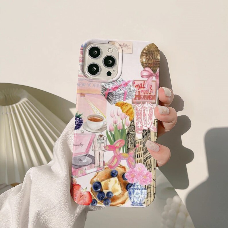 Op de afbeelding: Een witte iPhone-hoes met een kleurrijk collage-ontwerp. Het ontwerp bevat afbeeldingen van een skyline, een luchtballon, een platenspeler, een kopje koffie, een croissant en een bord met pannenkoeken. De tekst "mat" en "real heaven" is zichtbaar op de hoes.