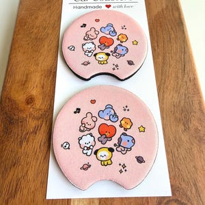 Könnte beinhalten: Zwei runde, rosafarbene Auto-Untersetzer mit bunten Zeichentrickfiguren, Sternen und Musiknoten. Die Präsentationskarte über den Untersetzern trägt die Aufschrift Car Coasters Handmade with love, mit einem roten Herzsymbol.