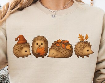 May include: A sand-colored sweatshirt featuring a row of four cartoon hedgehogs, each adorned with autumn-themed accessories like a hat and leaves. The sweatshirt is made of soft material and is labeled "Soft".