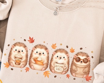 May include: Sand-colored sweatshirt featuring a charming illustration of four hedgehogs, each holding autumn-themed items like a pumpkin spice latte and a slice of pie. The phrase "Autumn Vibes" is on a mug. The sweatshirt is soft and made of Gildan 18000 material.